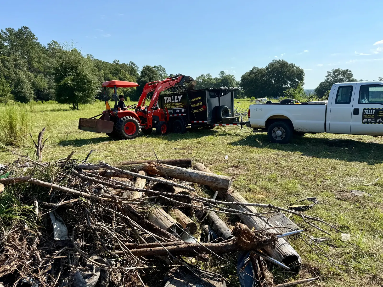 Kubota tractor with a grapple loading debris into the branded Tally Junk Removal trailer hitched to a white pickup