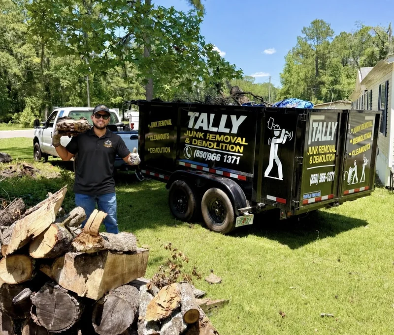 Tally Junk Removal owner loading cut logs into the branded black dump trailer in a Tallahassee yard