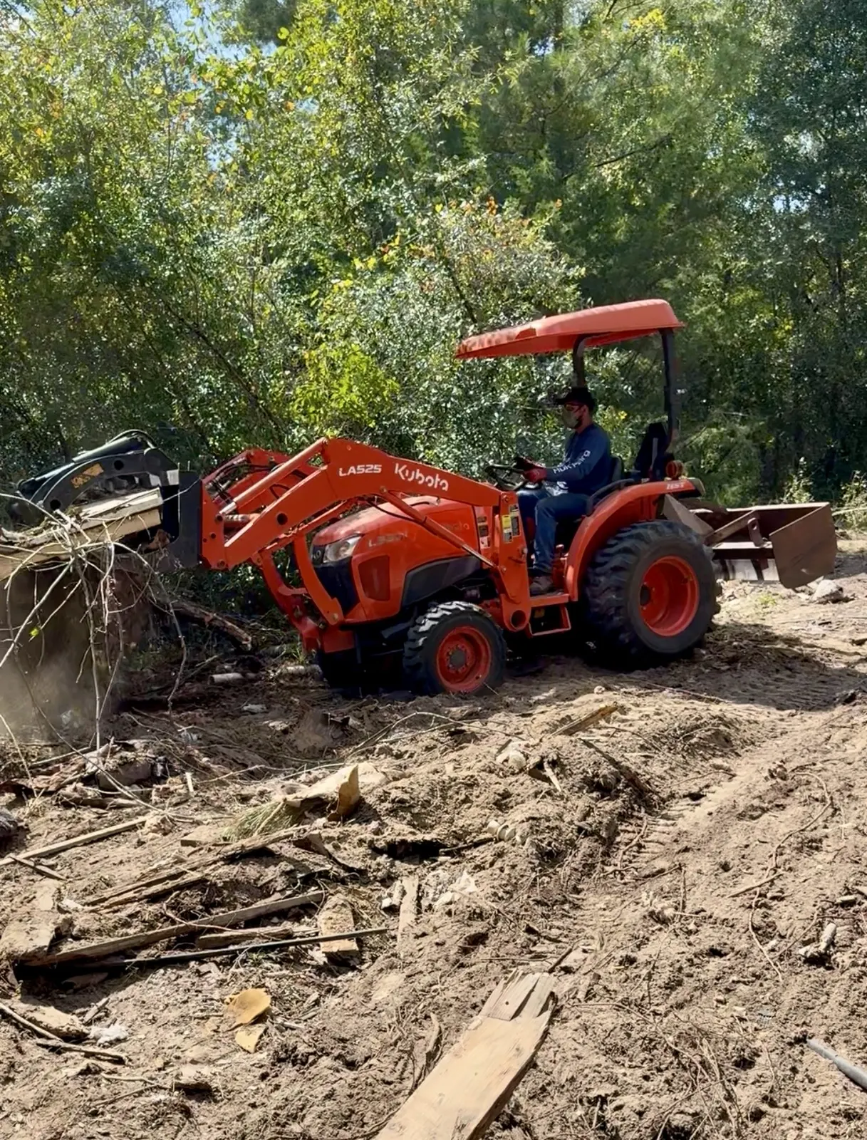 Tally Junk Removal owner operating a Kubota tractor with a grapple, loading debris on a wooded lot