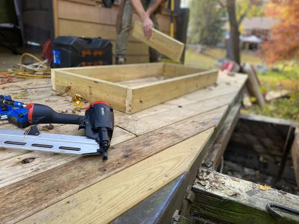 Wooden deck boards partially removed to expose the framing, with a nail gun resting on the decking