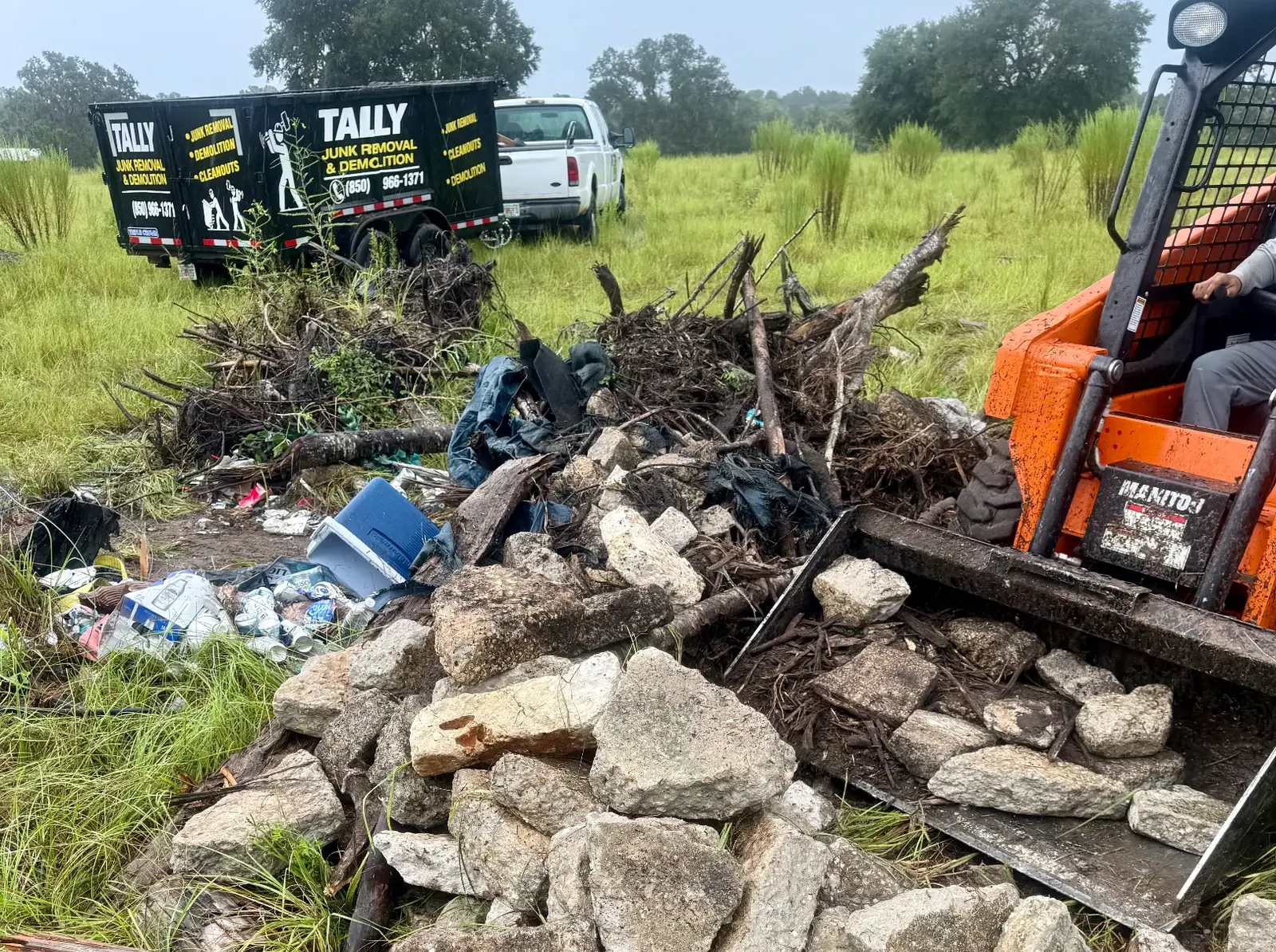 Skid steer bucket scooping concrete chunks beside piles of branches and trash, with the Tally trailer behind