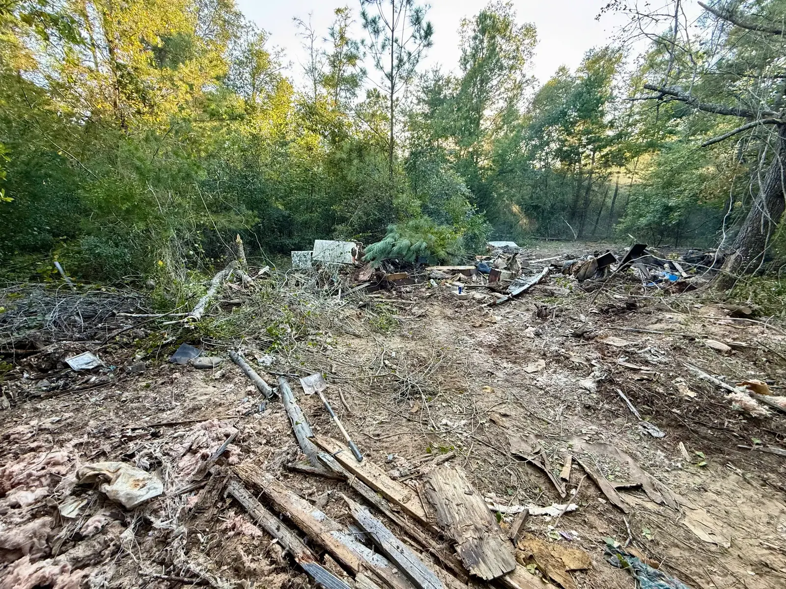 Wooded lot mid-cleanup with debris pushed into piles, loose insulation and lumber still on the ground, and a shovel