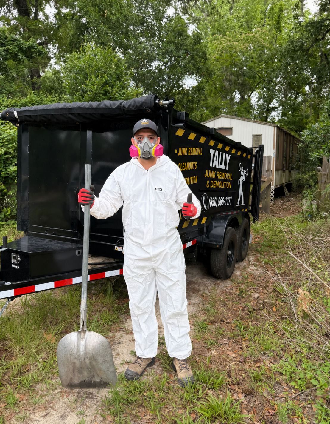 Tally Junk Removal team member in protective coveralls and respirator with a shovel next to the branded dump trailer