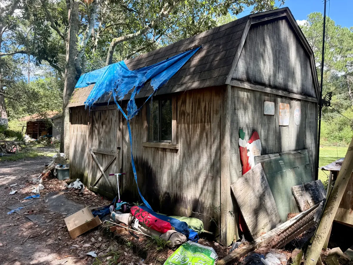 Deteriorating barn-style shed with a blue tarp on the roof before demolition