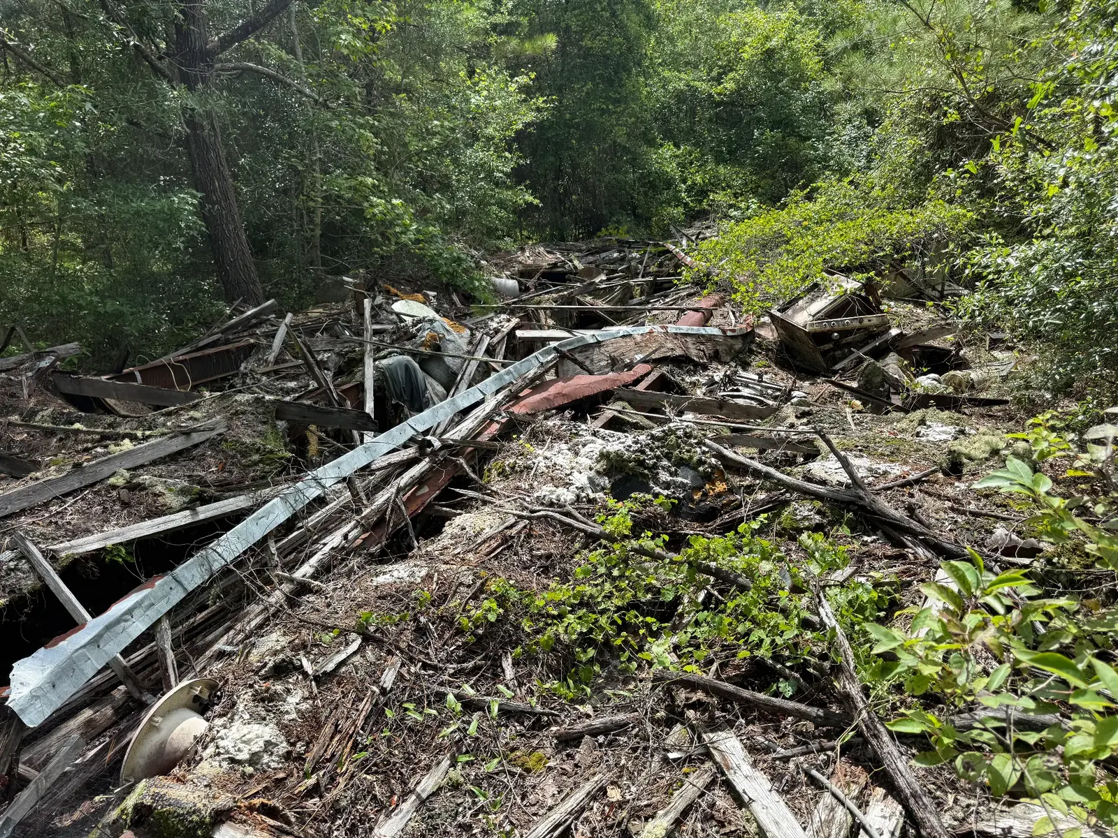 Overgrown collapsed debris on a wooded lot: metal trim, rotted lumber, steel frame rails, and an old stove