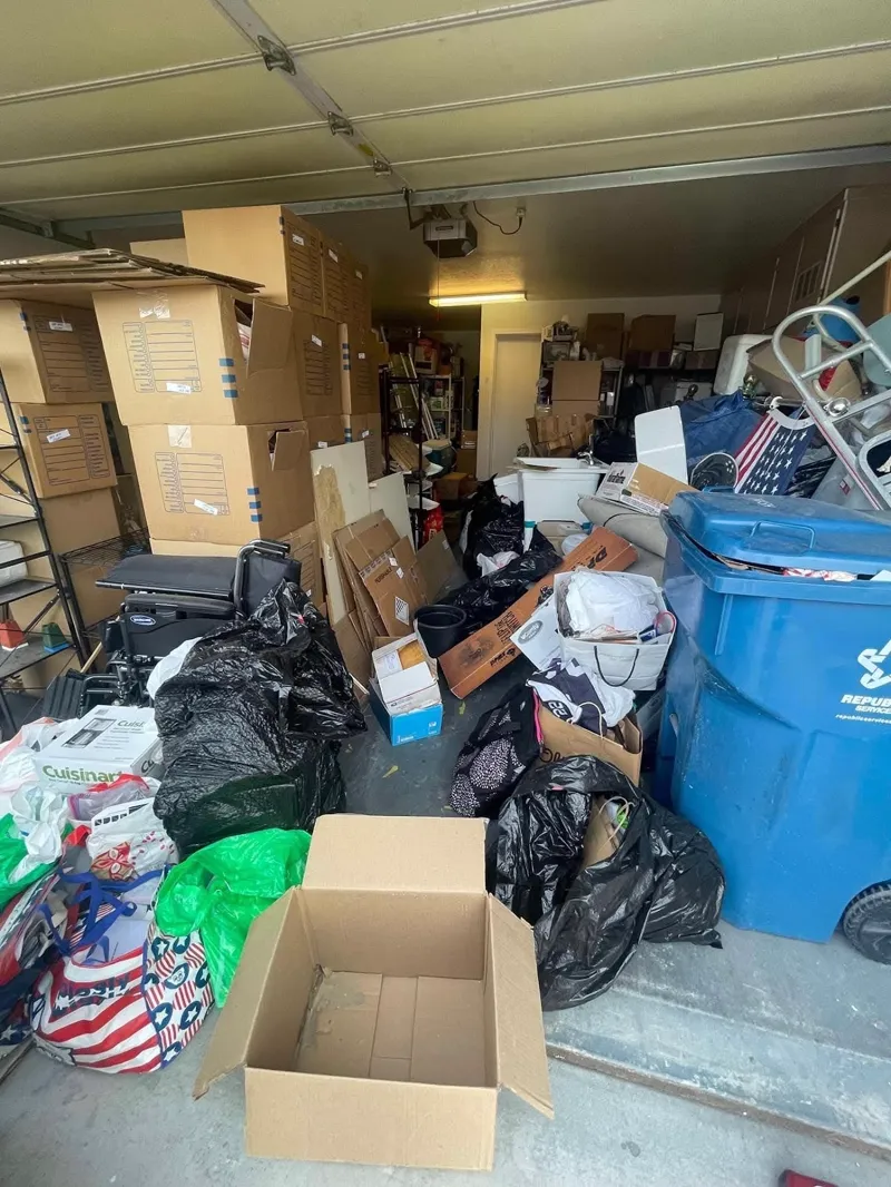 Garage filled wall to wall with stacked moving boxes, bagged items, a recycling cart, and a folded wheelchair