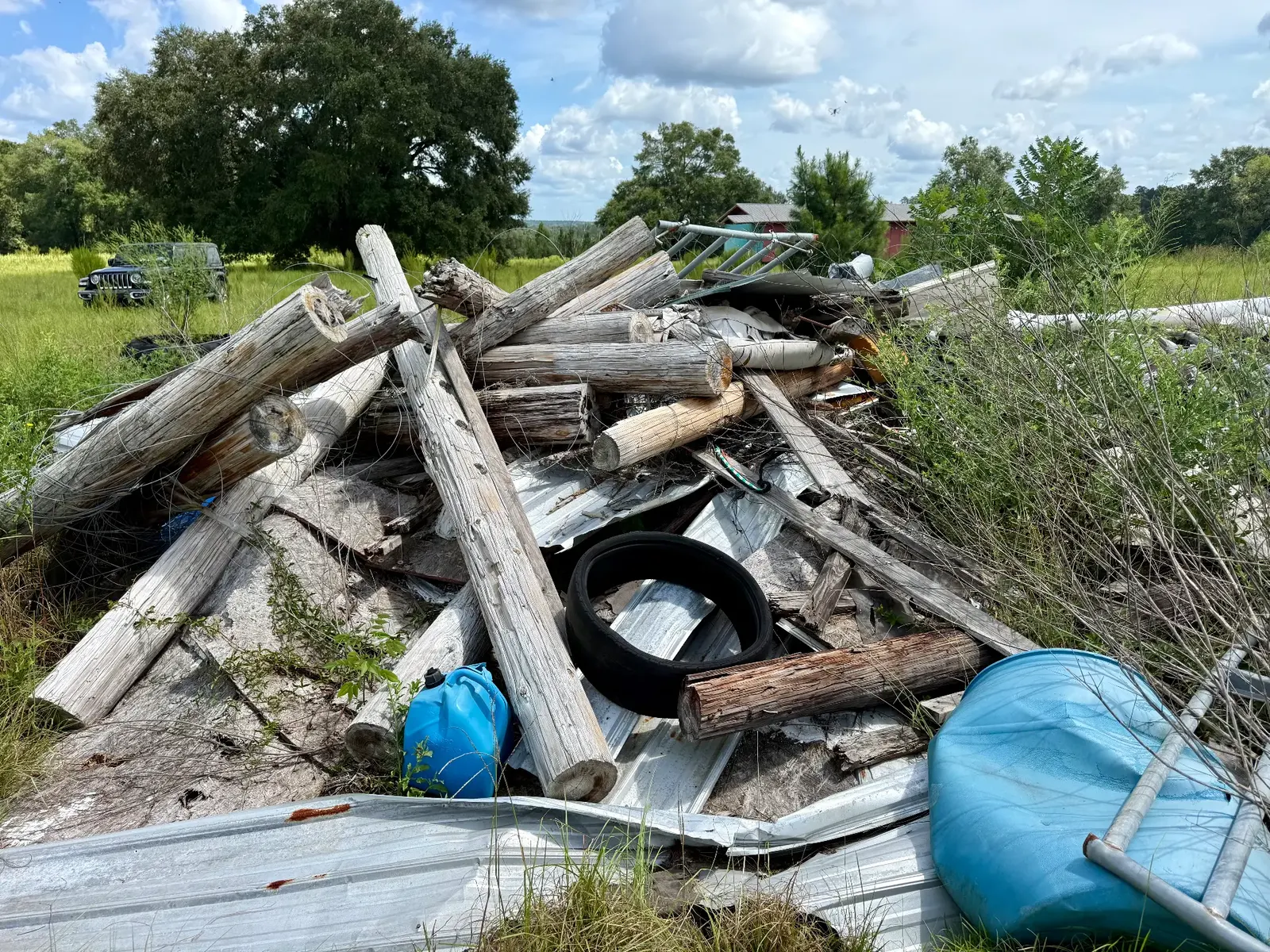 Large pile of old wooden poles, metal roofing, a tire, and plastic barrels in an overgrown field
